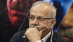 مردی میانسال با موی کم و عینک و لباس رسمی، دستش را روی دهان خود گذاشته و به سمت چپ نگاه می‌کند.