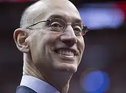A thin, bald man in a dark pinstripe suit, wearing glasses, striped shirt and dark blue tie against a gray background