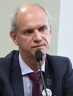 Middle-aged man with short hair speaking during a meeting. He is wearing a dark blue suit, with a white shirt and wine-colored neck tie.