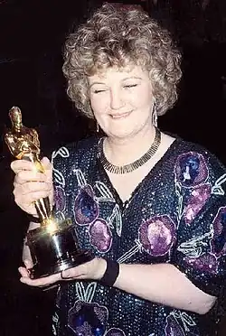 A picture of a curly-haired woman wearing a dress with various purple و green prints. She is holding a golden statuette.