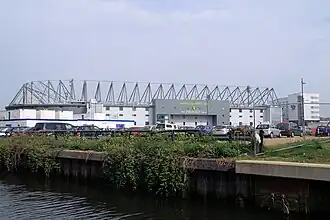 The exterior of Carrow Road, an association football stadium. A river is in the foreground.