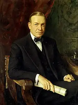 Man of about 65 in an ornate wooden chair. Formally dressed in a dark suit, white shirt, and black bow tie, he holds a rolled document in his left hand.