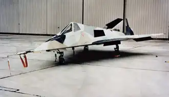 Small jet aircraft with angled surfaces in hangar. It is painted in a disruptive scheme to confused casual onlookers.