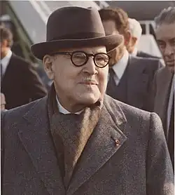 عکس یک مرد خندان را نشان می‌دهد که پالتو، عینک، کلاه و سبیل نازکی به‌صورت خود دارد.