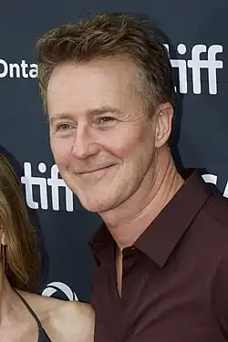 A portrait of Edward Norton, a blonde Caucasian man in a white plaid shirt. He is smiling towards the camera
