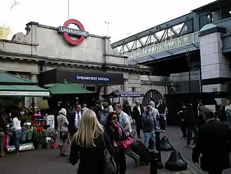 A grey building with a blue sign reading "EMBANKMENT STATION" in white letters and people standing in the foreground all under a white sky