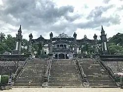 Staircase leading to a building of dark stone. A simple decorated gate is at the top of the staircase.