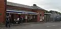 A brown-bricked building with a blue sign reading "EPPING STATION" in white letters and people in the foreground all under a blue sky with white clouds