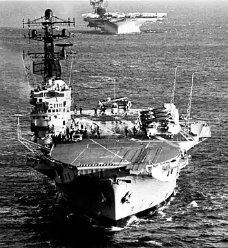 Bow view of an aircraft carrier sailing towards the photographer. Aircraft with folded wings are sitting on the deck. A second aircraft carrier is following the first, but the upper part of this ship is outside the frame of the photograph.