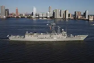 A frigate with the number "03" painted near the bow. The ship is travelling slowly, and white-uniformed people line the decks with a city skyline in the background.