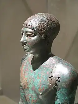 Closeup of the head of the statue of a man, made of greenish copper, its eyes inlaid in white and black stones