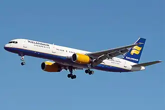 A mostly white Boeing 757 with blue and yellow trim preparing for landing against a blue sky. Landing gear and flaps are fully extended in final approach configuration.