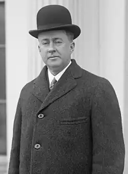 Cleanshaven man with drooping eyelids, aged about 40. He is wearing a black bowler hat, white shirt, tie and dark overcoat.