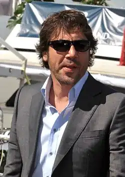 A picture of a Spanish man with sunglasses. He is wearing a greay coat over an unbuttoned light blue collared shirt.