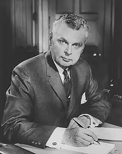 Half-length photo portrait of a man aged about 60. He has a square face, strong jaw, firm lips and piercing pale-coloured eyes under thick arched eyebrows. His full head of curly hair is streaked with grey, cut short and combed back smoothly with hair oil. He wears a neat suit and tie, and holds a document, looking at the camera with a penetrating stare.