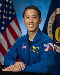 An Asian man in a blue flight suit is sitting in front of US & NASA flags; he is facing the camera and smiling.
