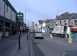 Street scene showing shops on left and right, with cars and vans on road. On the left hand pavement is a sign saying welcome to Keynsham high street.