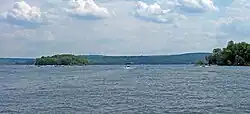 Image of a boat moving away from the photographer on a wide lake with two forested islands visible in the foreground. Mountains rise in the background.