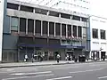 A grey building with blue panels and a dark blue, rectangular sign reading "LANCASTER GATE STATION" in white letters and people in front