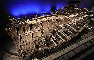 A diagonal section of a wooden ship seen from the stern inside a moodily lit building while it is being sprayed with water from a sprinkler system