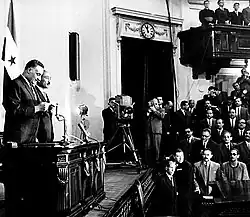 Two men on a stage, with a flag hung behind them. One is reading from a paper, while the other is looking at the audience. Cameras are shooting the event, while most of the audience is looking at the stage.