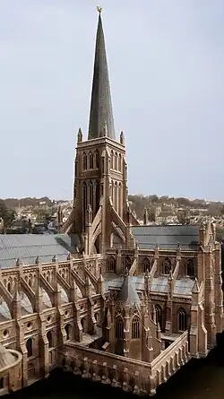 The central tower of a large gothic cathedral. The central tower is buttressed and with an imposing wooden spire. An octagonal chapter house is in the foreground.