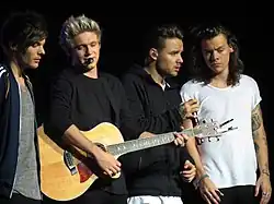 Four men standing on stage, the second man from the left holding a guitar