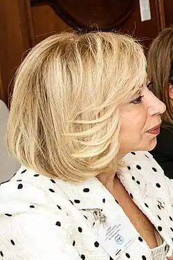 An official portrait of Neemat Aoun, a middle-aged woman with blonde hair styled in an elegant bob, dressed in a sophisticated black-and-white suit.