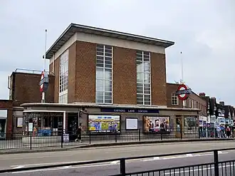 A brown-bricked building with a rectangular, dark blue sign reading "RAYNERS LANE STATION" in white letters all under a dark blue sky