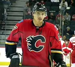 A hockey player with short dark hair looks to his right as he skates. He is in a white uniform with red and blue trim, the number 27 on his arms and a stylized "CH" logo on his chest.