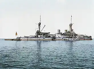 A large light gray battleship with two tall masts sits motionless in calm water. Several smaller boats are tied alongside.
