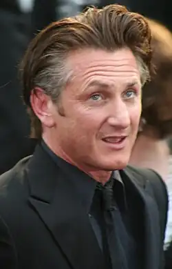A picture of a man with wavy brown and grey hair is seen wearing a black suit, shirt, tie, and coat.