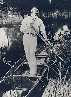 Paget standing in a canoe wearing a deerstalker cap, holding an oar