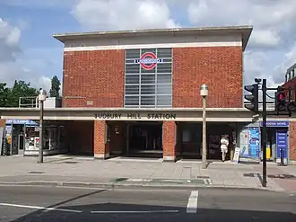 A red-bricked building with a rectangular, grey sign reading "SUDBURY HILL STATION" in black letters all und er a blue sky with white clouds