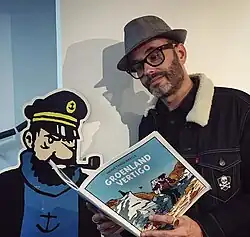 عکسی از یک مرد با عینک و کلاه، در حال خواندن کتاب کمیکی که در دست دارد، در مقابل یک برش مقوایی از کاپیتان.