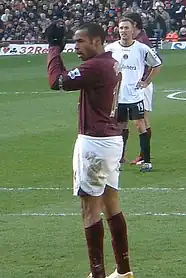 Freddie Ljunberg, wearing gloves and a redcurrant football shirt applauds the crowd. A stand full with people and man wearing a football shirt are visible in the background.