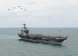 A modern aircraft carrier at sea with five aircraft flying in formation overhead