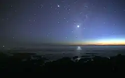 A photograph of the night sky taken from the seashore. Many glimmers of sunlight is on the horizon. There are many stars visible. Venus is at the center, much brighter than any of the stars, and its light can be seen reflected in the ocean.