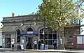 A beige-bricked building with a rectangular, dark blue sign reading "WEST BROMPTON STATION" in white letters all under a light blue sky