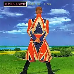 A man looking out to the plains with his back on the view. He is seen wearing a coat with the Union Jack on it in a poor condition.