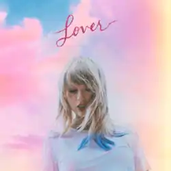 A picture of Swift wearing a blue shirt and blue-highlighted blonde hair with a glitter heart shape on her right eye, looking downward. The background features a cloudy sky filled with blue, pink, and yellow hues. The title "Lover" is written in a red cursive font at the upper corner.