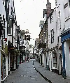 Street scene. Buildings lining narrow lane with central water gully, pedestrians, and hanging baskets.