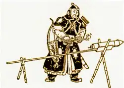 مردی با زره سیاه ایستاده در مقابل یک راکت که به یک چوب متصل شده‌است و آن چوب توسط دو پایهٔ چوبی به شکل X نگه داشته شده‌است.