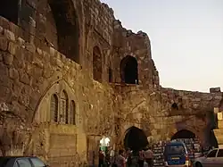خودروها و مردم در مقابل یک دیوار سنگی با دهانه‌های پنجره و در و بقایای طاق‌ها