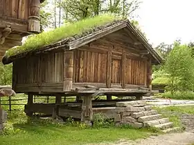 Crude log and timber open-air cabin with a pitched roof on a foundation of rocks, with the second storey overhanging the first.
