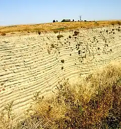  This photo shows a canyon cut into the surrounding flat soil with 32 distinct horizontal layers of soil, each clearly demarked from the layer below. Above the canyon a farm house can be seen in the distance - the farm house provide the perspective that helps the viewer establish that the cut is over 40 deep. The bottom of the cut is filled with tumble weeds.