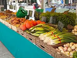A stall at an outdoor market displaying pumpkins, sweet potatoes, onions, corn, herbs, and other vegetables arranged in wooden crates.